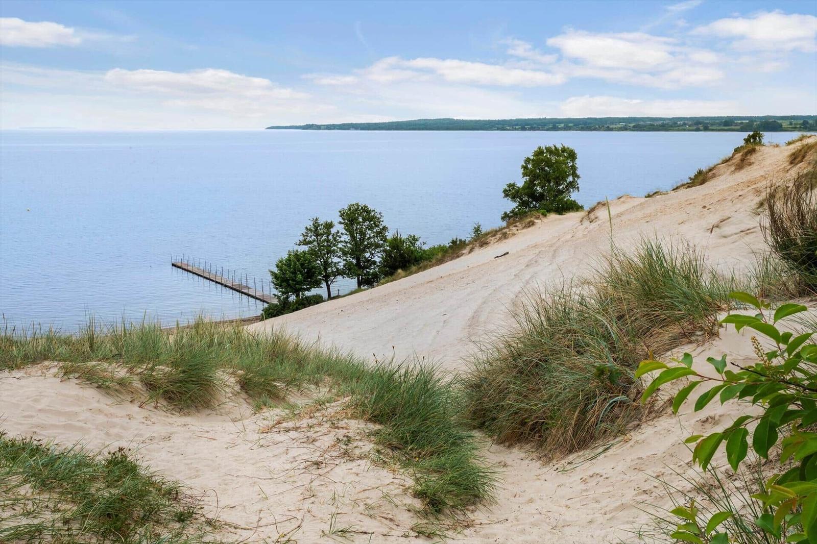 Sandhügel mit Gras, Blick auf ruhiges Wasser und einen Steg.