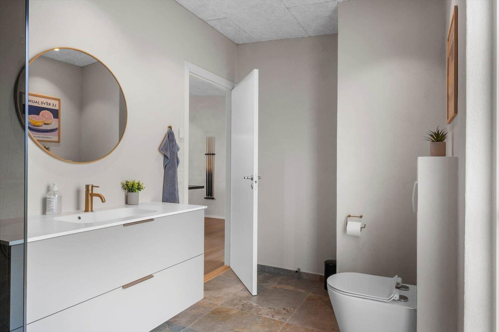 Badezimmer mit Waschbecken, Spiegel und Toilette. Tür führt in angrenzenden Raum.