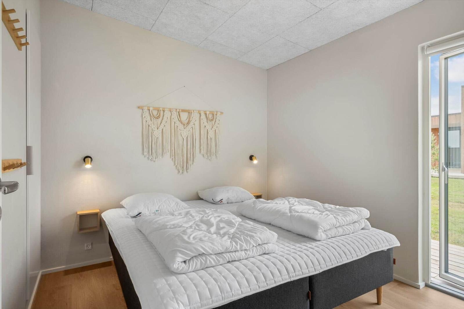 Schlafzimmer mit Doppelbett, weißen Bettwäsche und Holzfußboden. An der Wand hängt ein Macramé-Teppich.