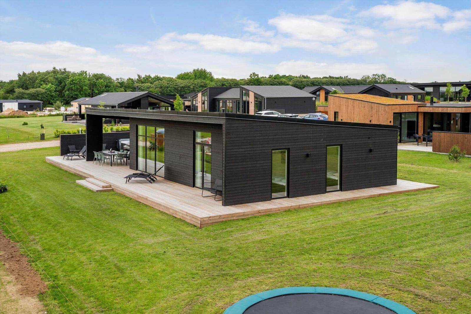 Moderne Villa mit Holzterrasse und Garten. Mehrere Gebäude im Hintergrund.