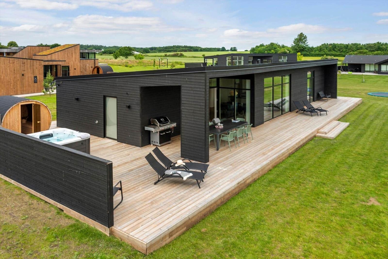 Moderne Villa mit großem Holzdeck, Whirlpool, Grill und Loungebereich. Blick auf grüne Landschaft.