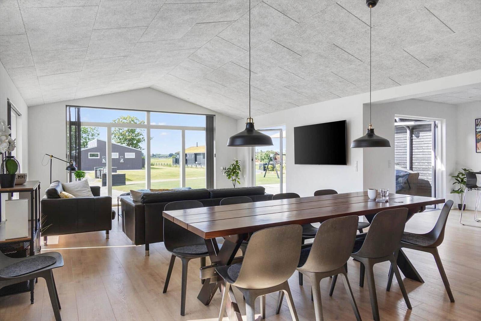 Dining area with wooden table and chairs. In the background, a living area with sofa and large window.
