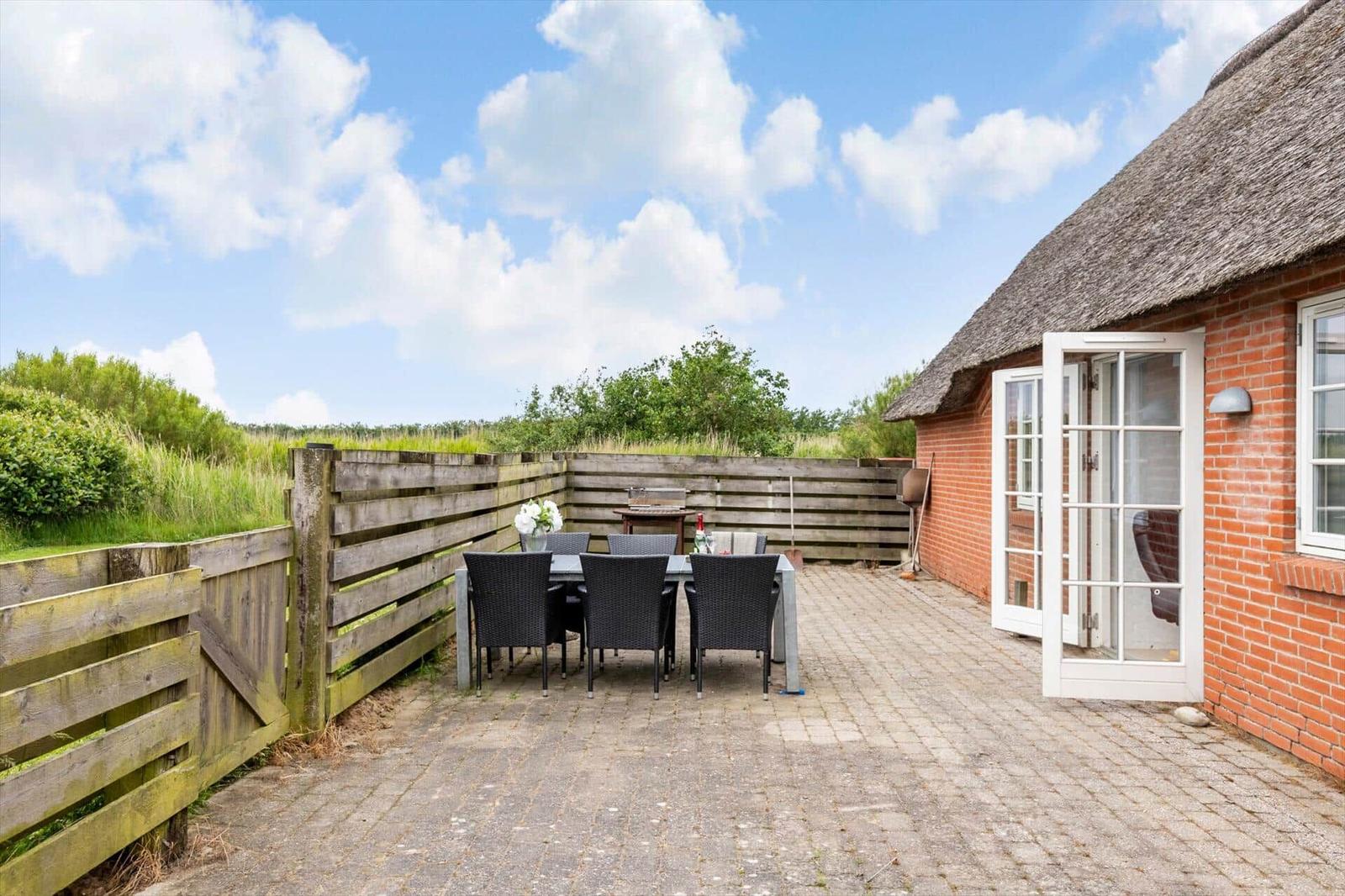 Terrasse mit Tisch und Stühlen neben einem Haus mit Strohdach und Holzumzäunung.