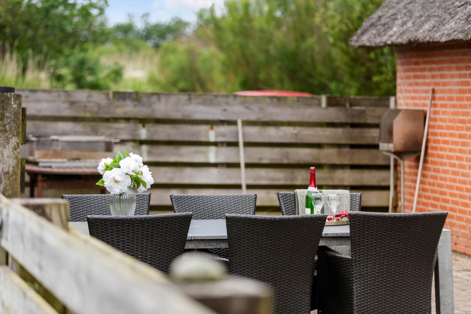 Terrasse mit Tisch, Stühlen, Blumen und Weinflasche. Hintergrund: Holzzaun und Backsteinhaus.