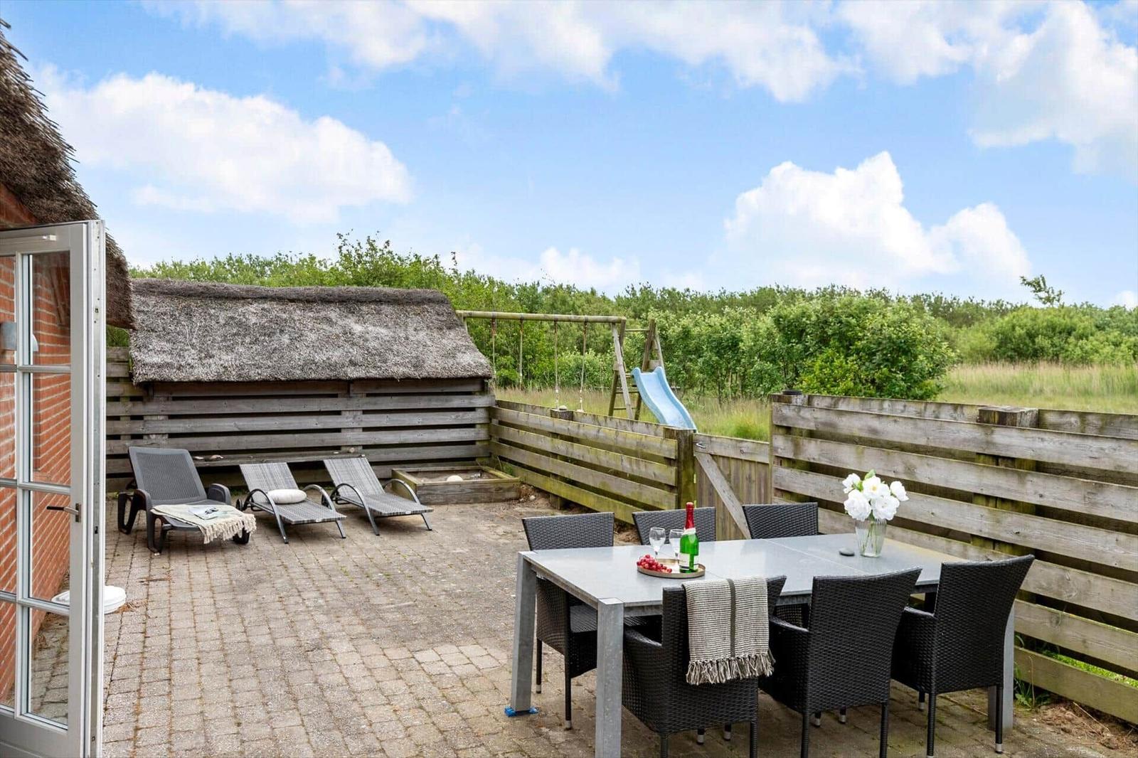 Terrasse mit Tisch, Stühlen, Liegen und Spielplatz. Hinterhof mit Holzzaun und Grünfläche.