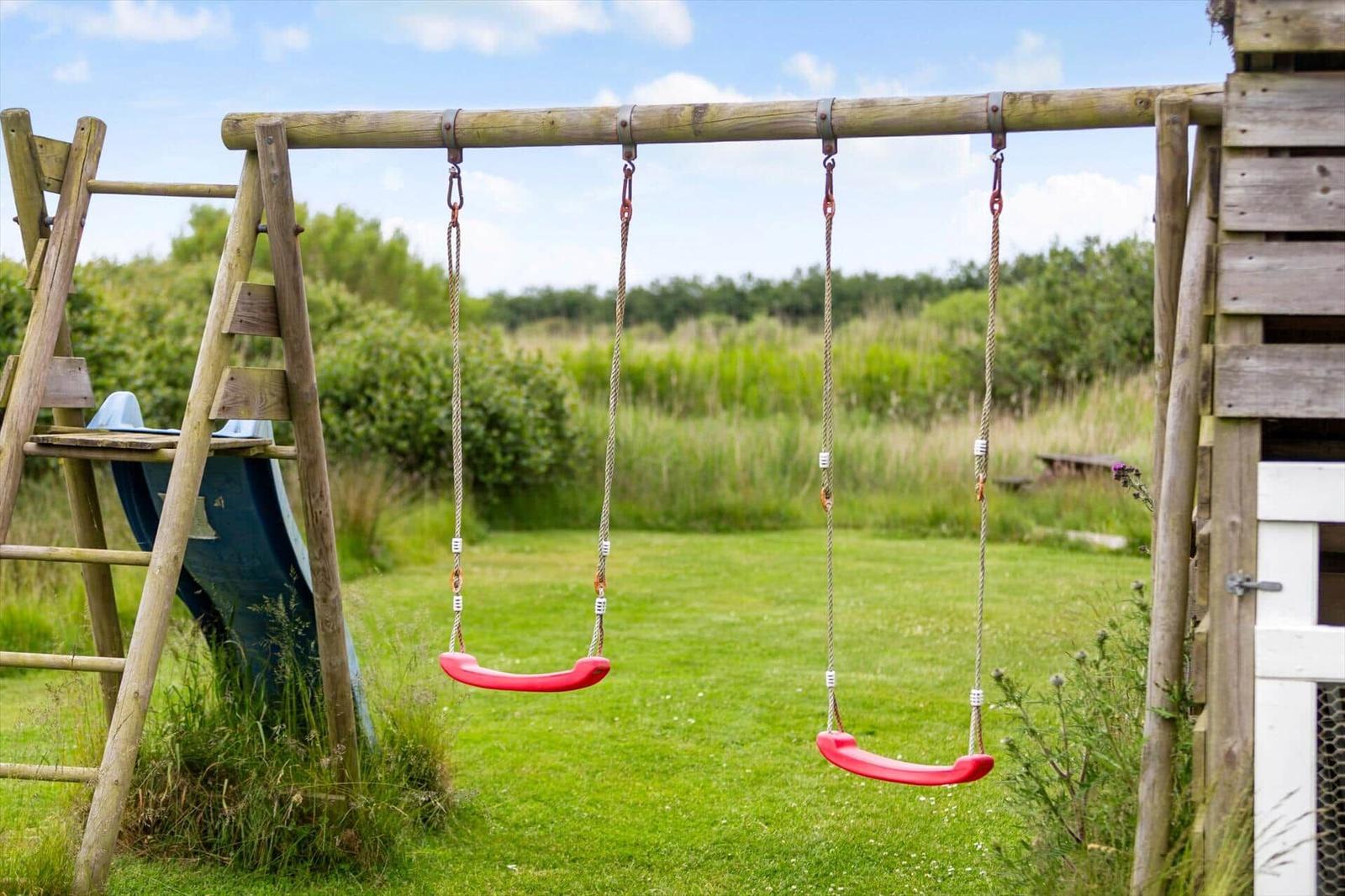 Zwei rote Schaukeln hängen an einem Holzgestell im Garten neben einer Rutsche.