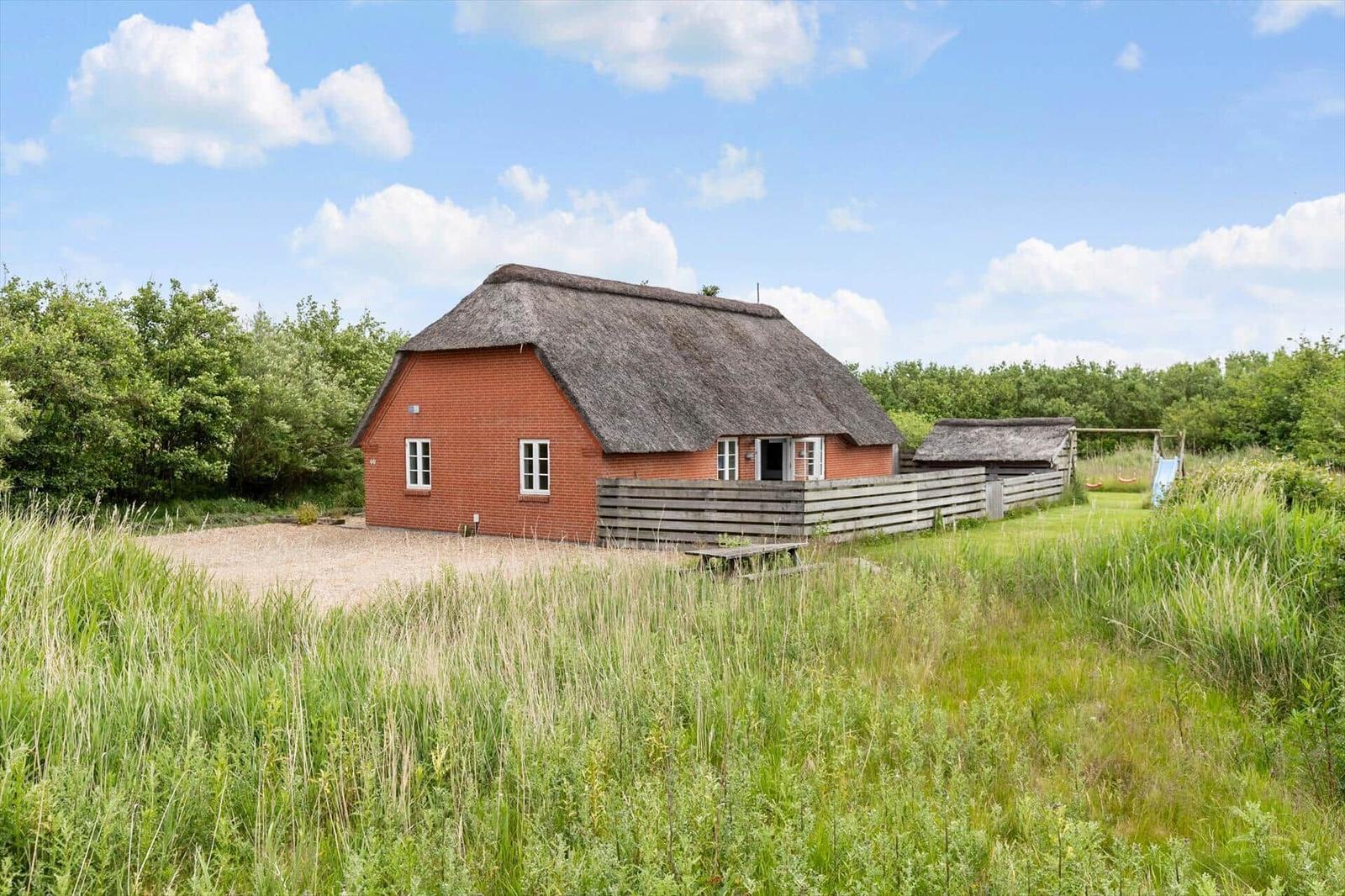 Rotes Haus mit Strohdach, umgeben von Gras und Bäumen unter blauem Himmel.