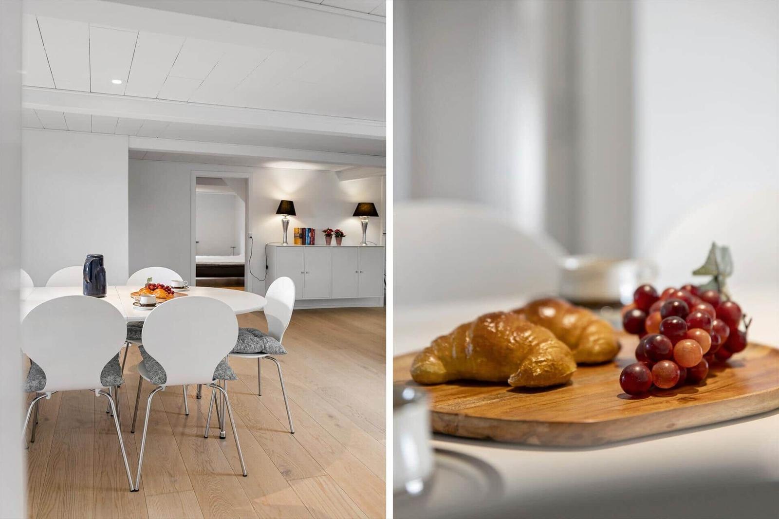Dining room with white table and chairs. Breakfast board with croissants and grapes.