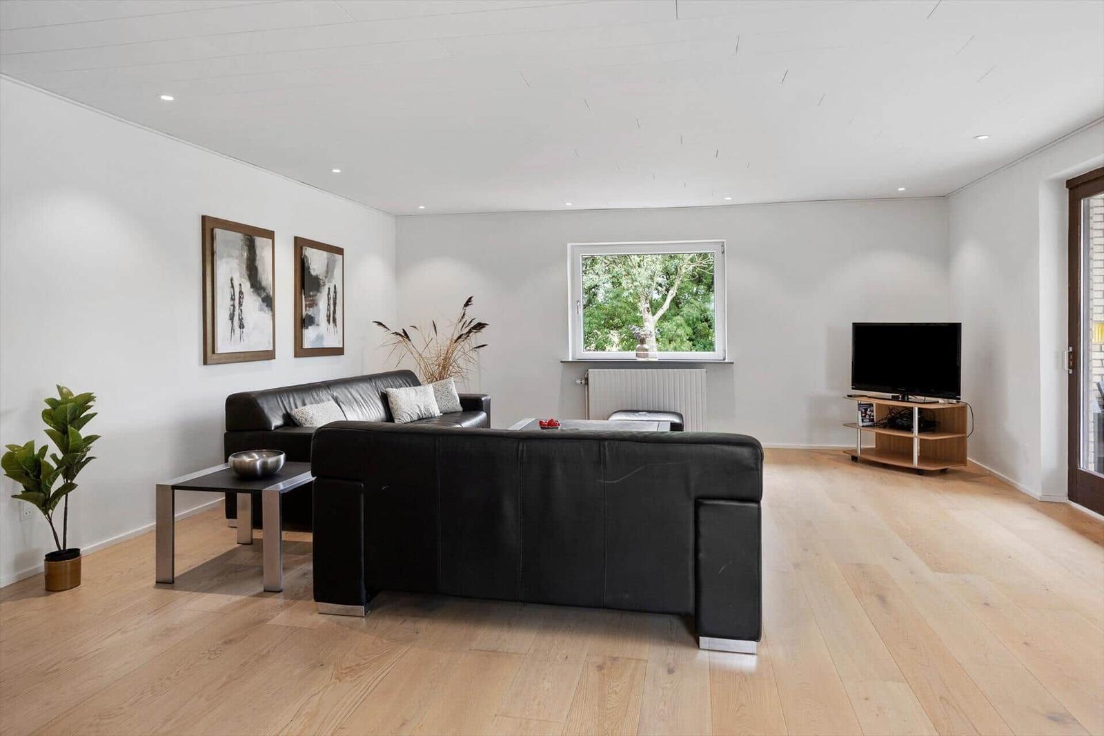 Living room with black leather sofa, wooden floor, and window with view of trees.