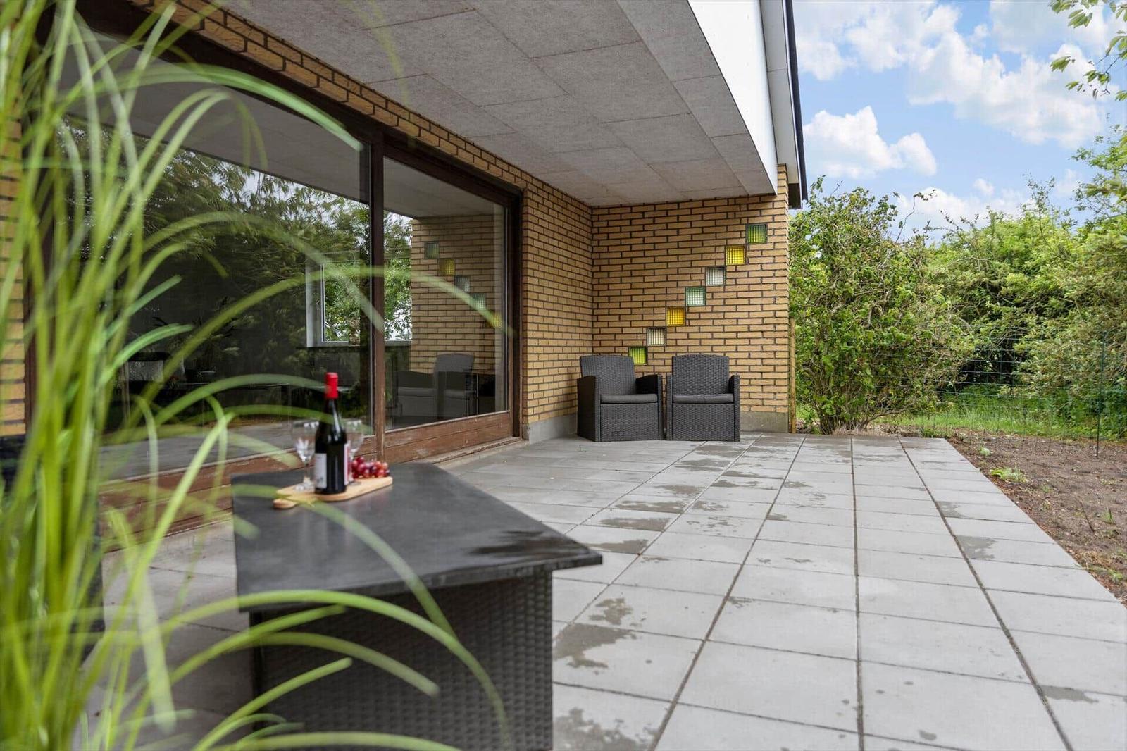 Terrace with seating and table. Background: house wall with glass tiles and green areas.