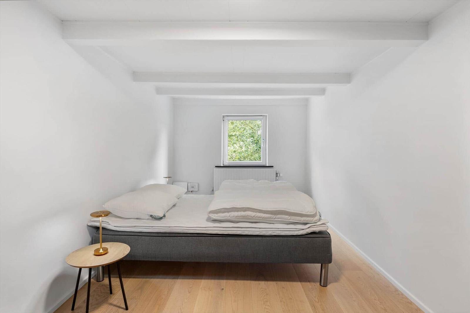 Bedroom with bed, small table, and window. Wooden floor and white walls.