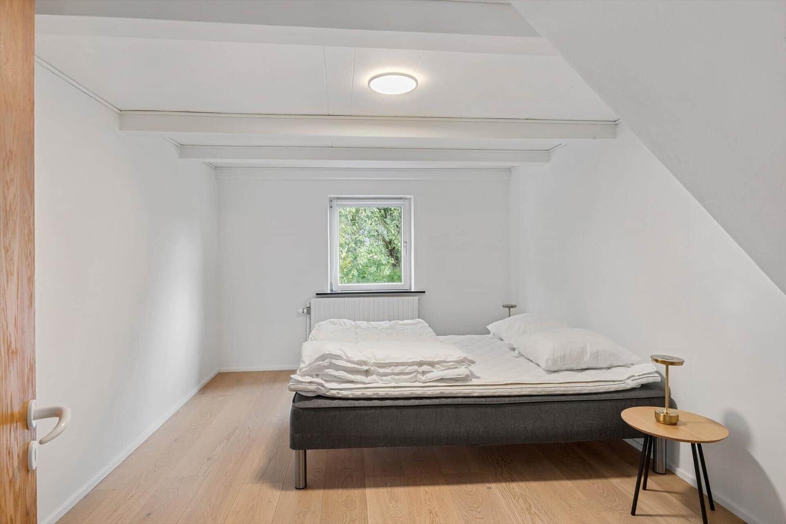 Bedroom with sloped ceiling, bed, side table, and window with view of trees.