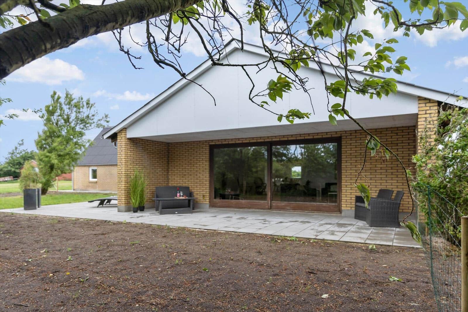 Modern villa with terrace, seating, and glass front. View of garden and sky.