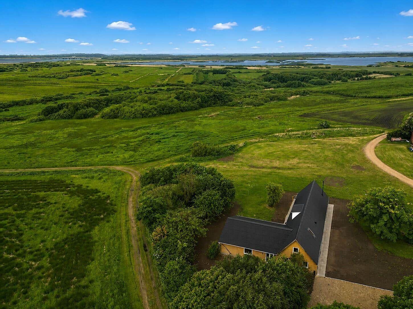 A house with a black roof is situated in a green landscape with meadows and a lake in the background.