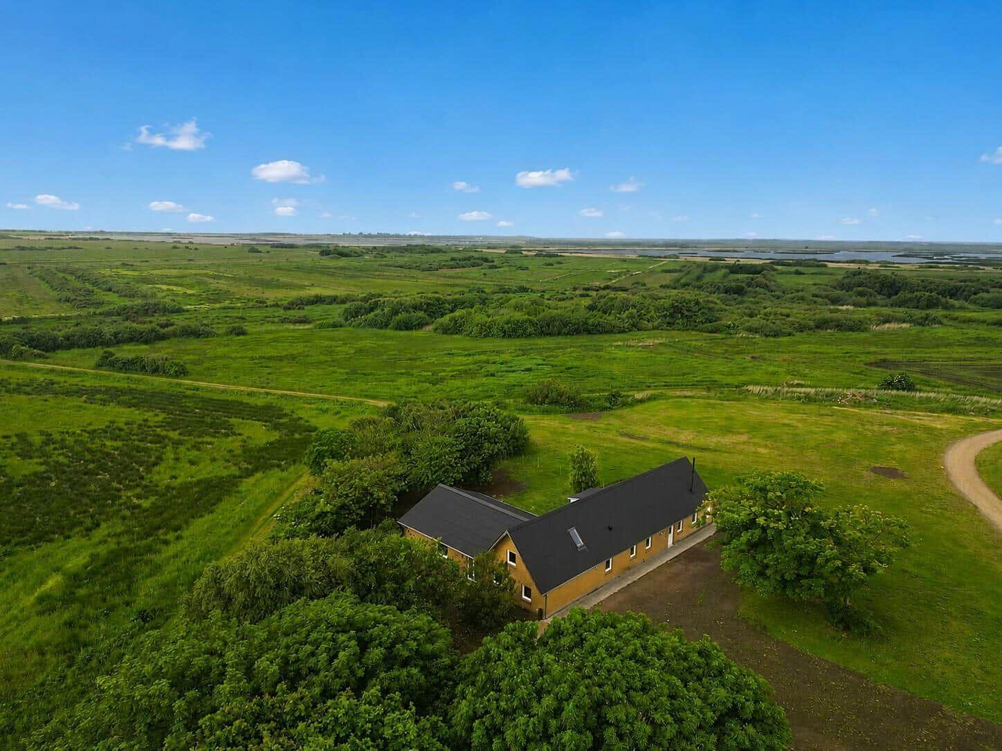 A yellow house with a black roof is situated in a green landscape with meadows and trees.