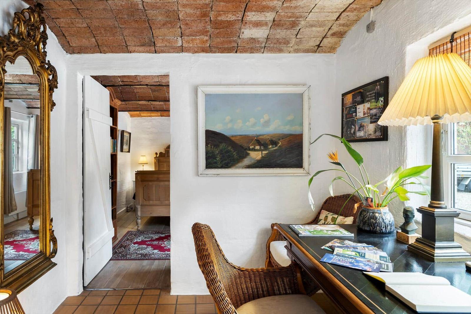 Raum mit Schreibtisch, Wickerstühlen und Bild an der Wand. Holzdecke und offener Durchgang.