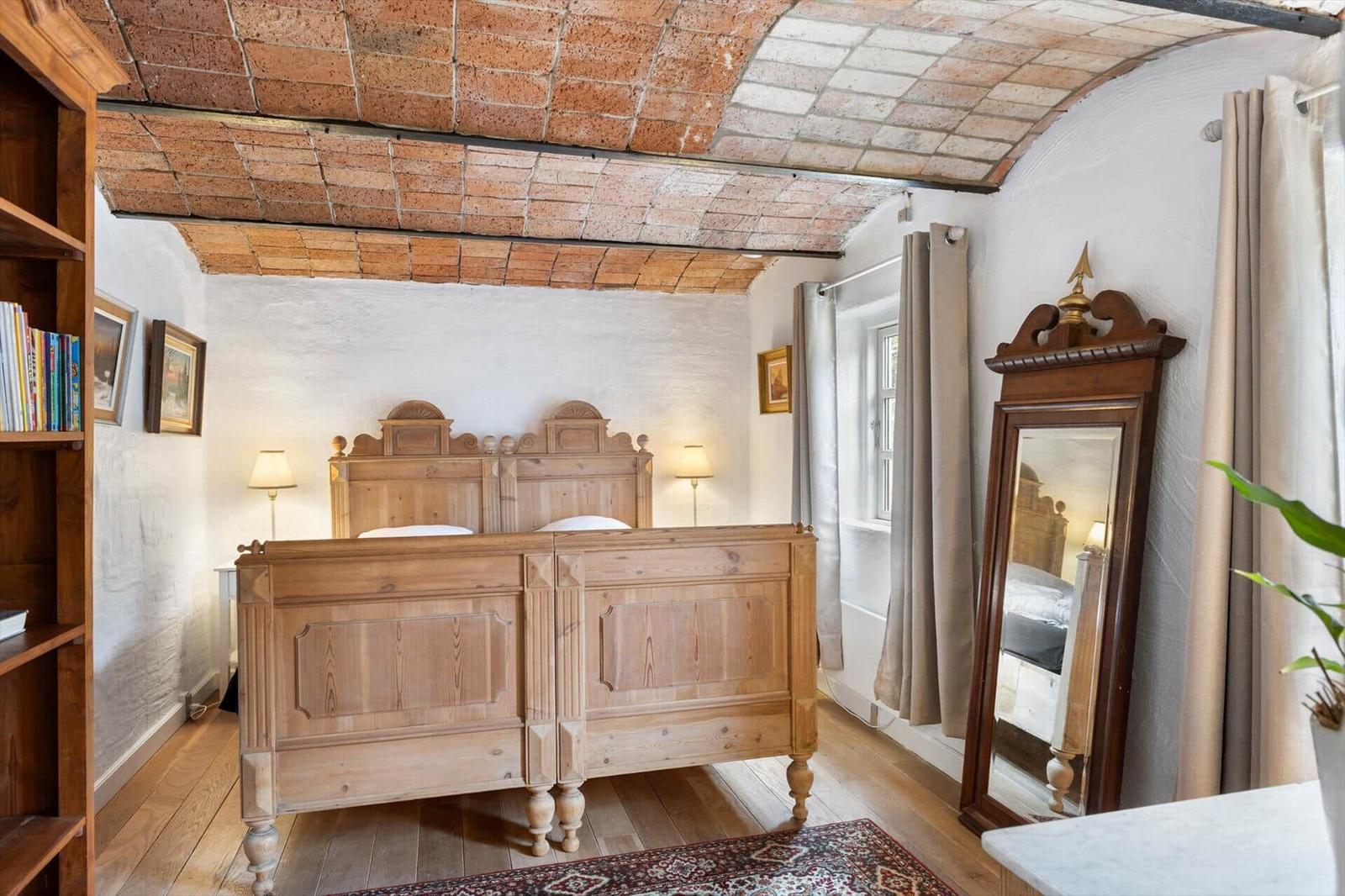 Schlafzimmer mit Holzbett, Steindecke und Spiegel. Holzfußboden und Bücherregal sichtbar.