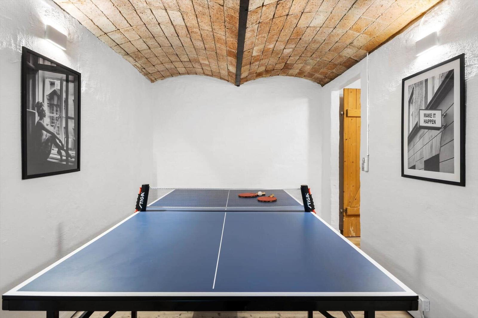 Tischtennisplatte mit Netz und Schlägern in einem Raum mit Steinbogendecke und Wandbildern.