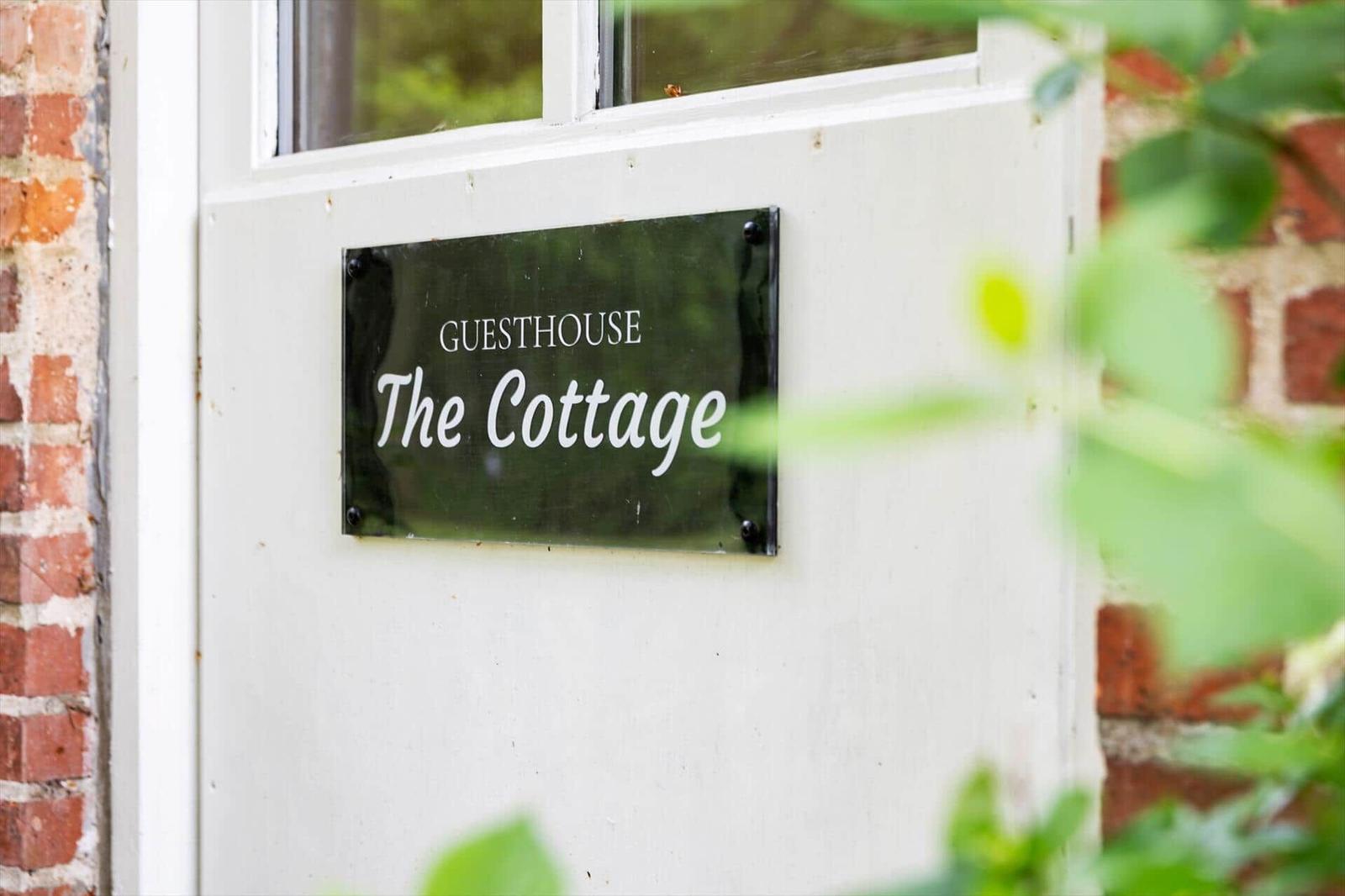 Schild an der Tür: Gasthaus The Cottage. Weiße Tür, rote Ziegelwand, grüne Pflanzen im Vordergrund.