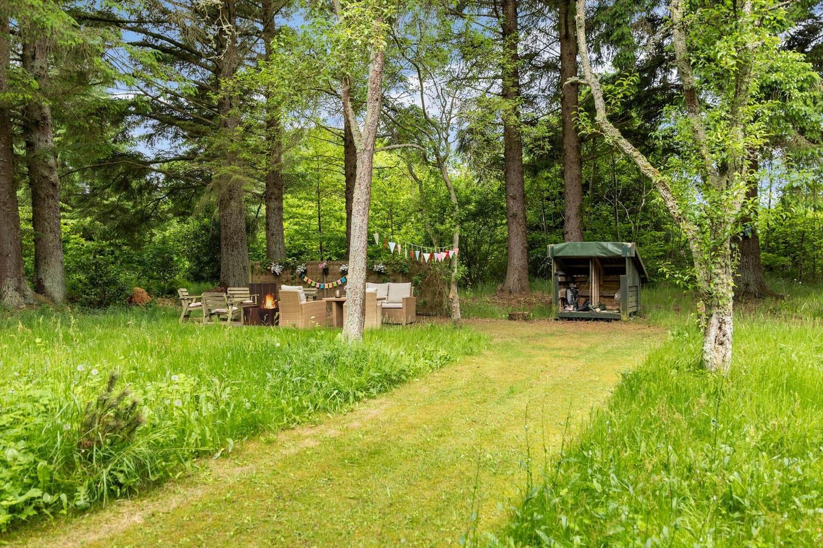 Grüner Garten mit Sitzbereich, Feuerkorb und Hütte zwischen Bäumen.