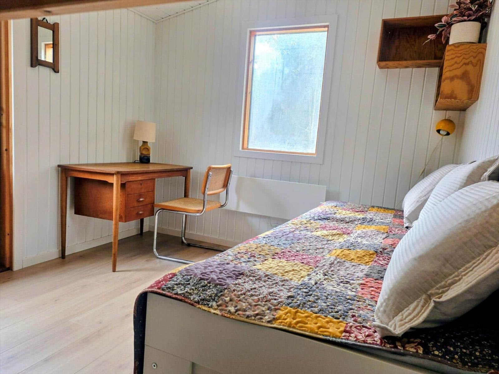 Schlafzimmer mit Bett, Schreibtisch und Fenster. Wandverkleidung und Holzboden.