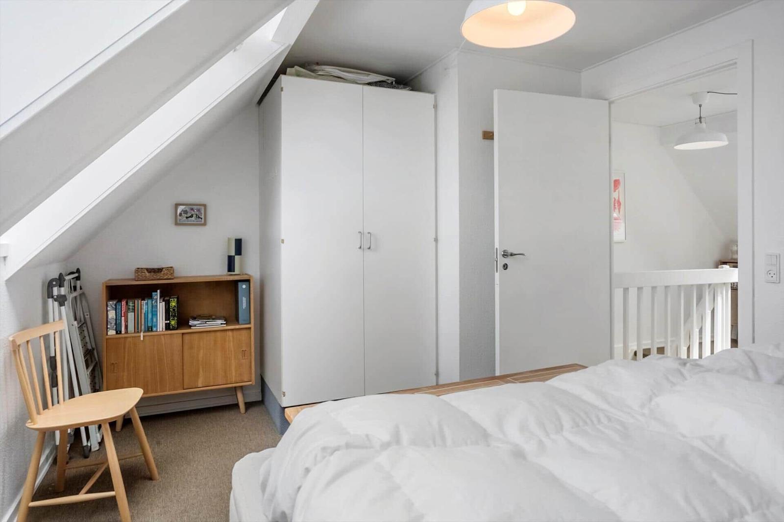 Schlafzimmer mit Dachschrägen, weißer Kleiderschrank, Holzregal und Bett mit weißer Bettwäsche.