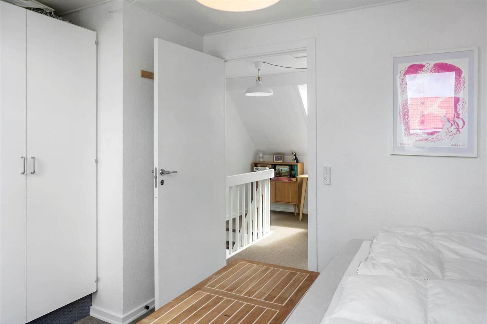 Schlafzimmer mit weißem Schrank, Bett und Tür zu einem Flur mit Holzboden und weißer Brüstung.