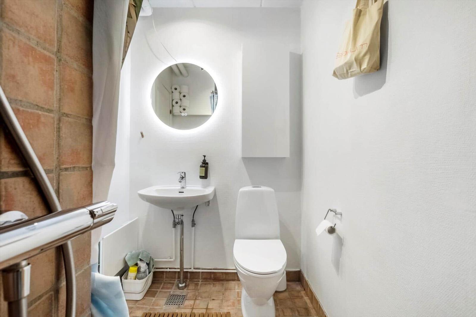 Badezimmer mit Toilette, Waschbecken und runden Spiegel mit Beleuchtung.
