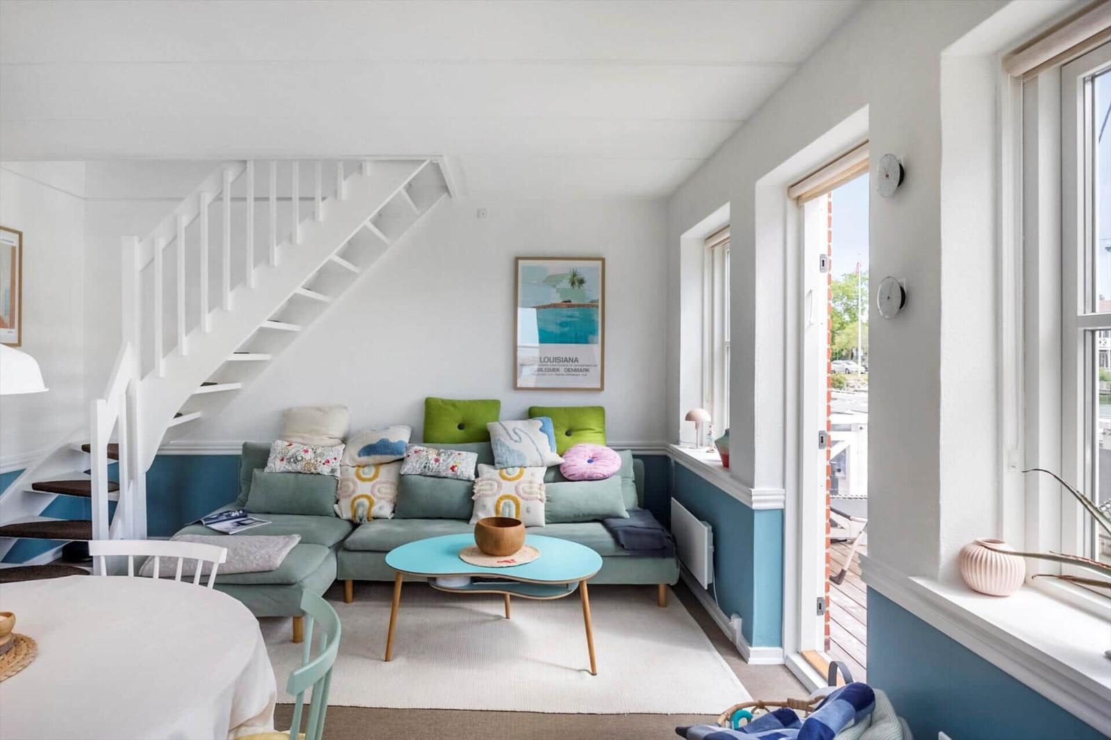 Wohnzimmer mit Sofa, Tisch und Treppe. Wandfarbe weiß und blau. Fenster mit Blick nach draußen.