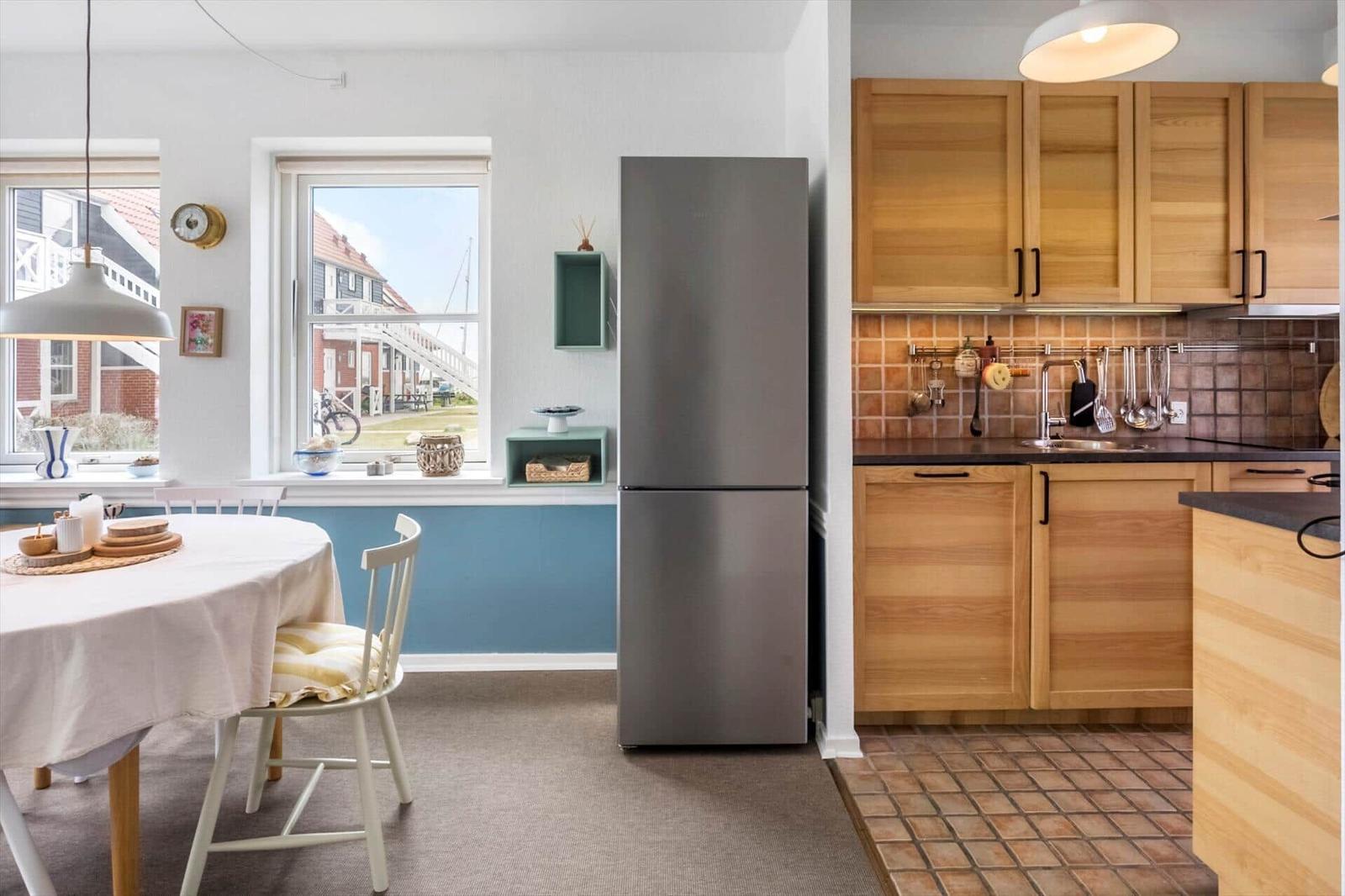 Küche mit Holzschränken und Kühlschrank. Essbereich mit Tisch und Stühlen. Fenster mit Blick auf Außenbereich.