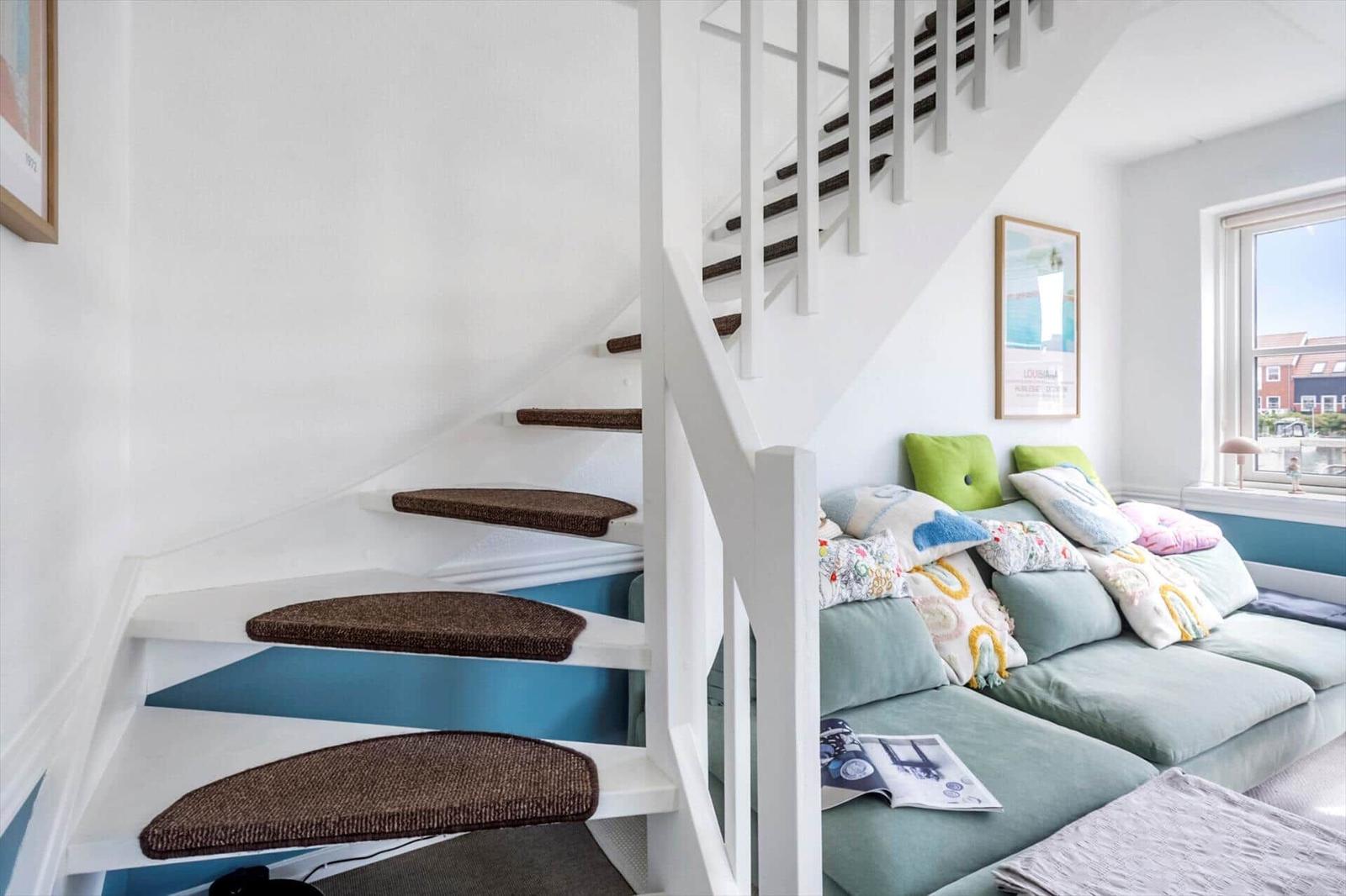 Eine Treppe mit braunen Stufen führt zu einem Wohnbereich mit Sofa und Fenster.