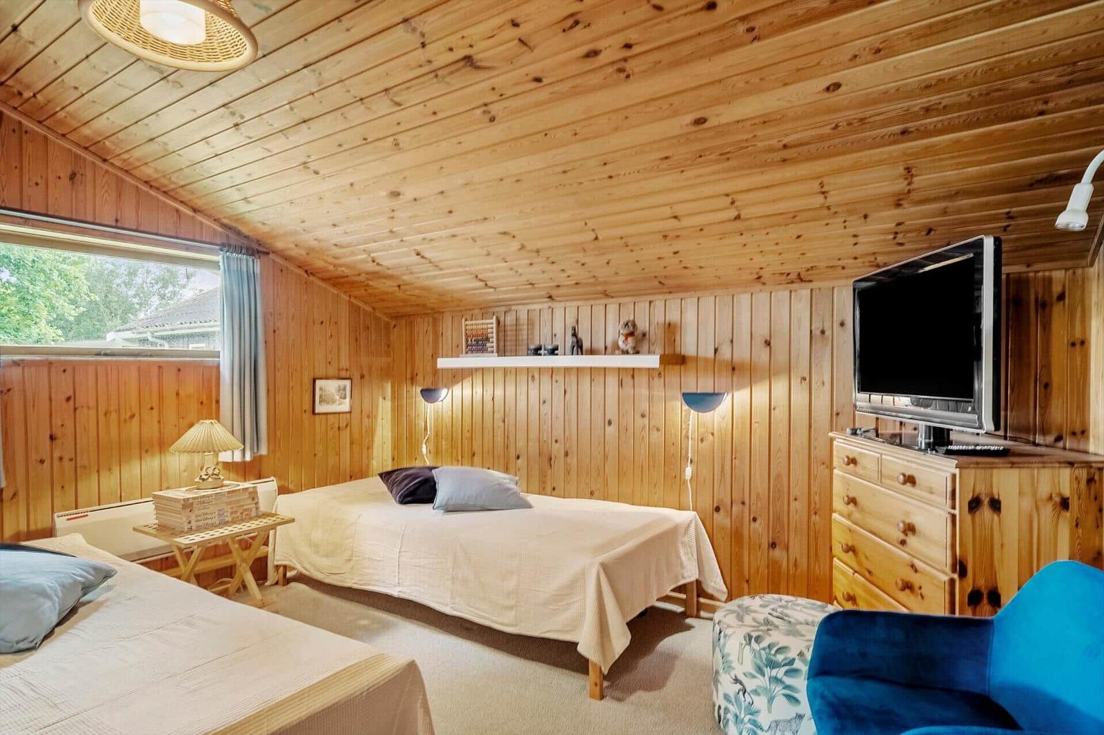 Zimmer mit Holzwand und -decke, zwei Betten, Fernseher und blauer Sessel.
