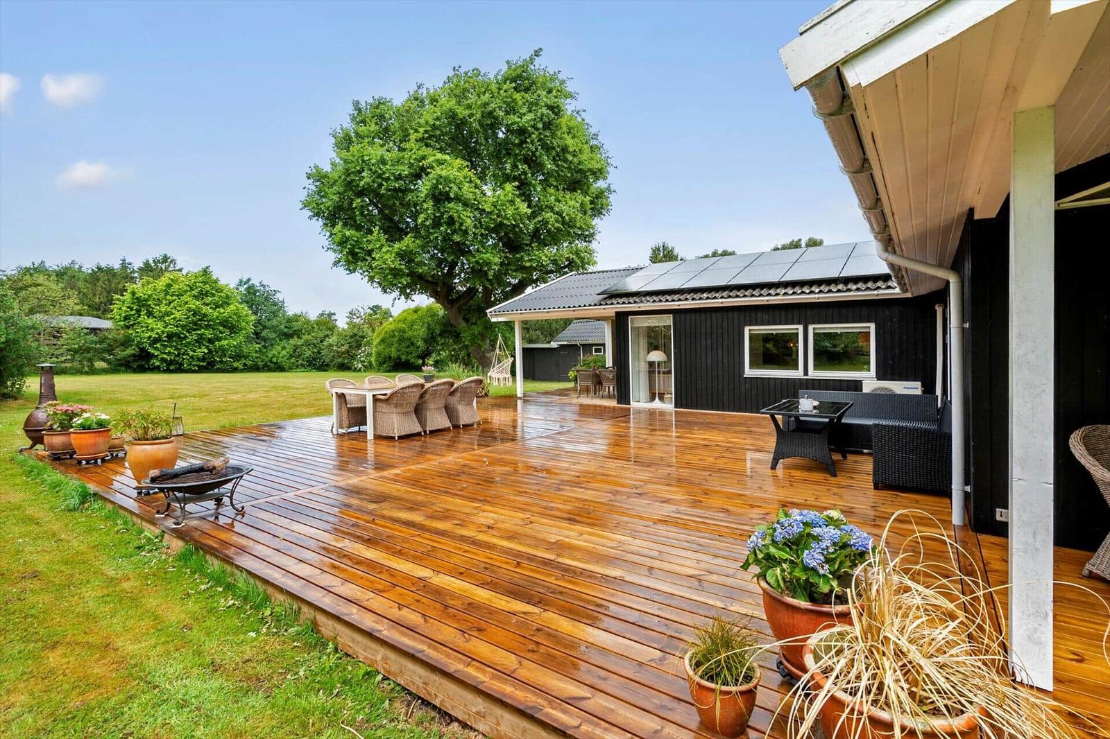 Holzterrasse mit Sitzgruppe, Garten und Baum. Schwarzes Haus mit Terrassentüren.