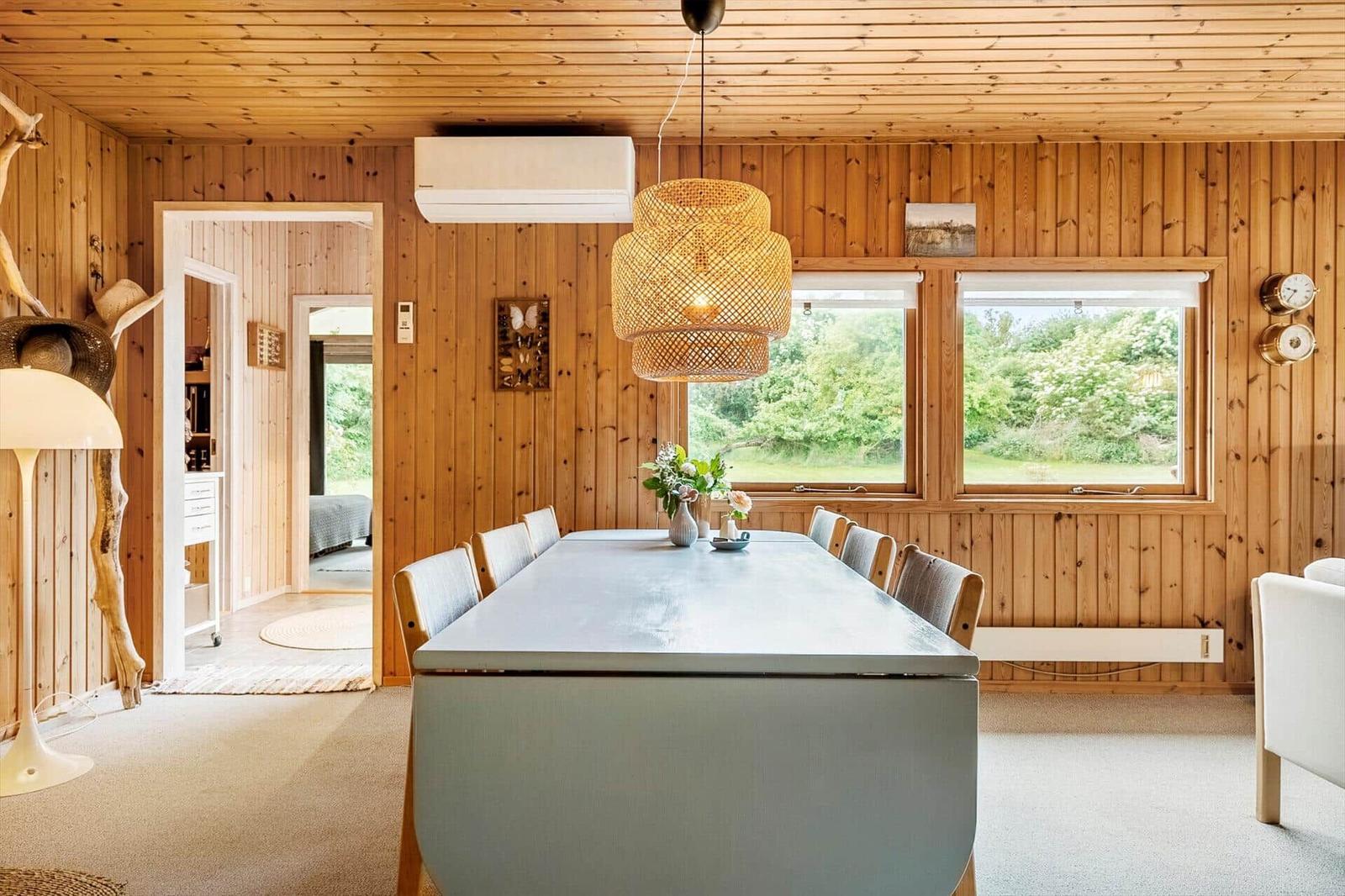Esszimmer mit langer Tisch, Holzwand und Fenstern zum Garten.