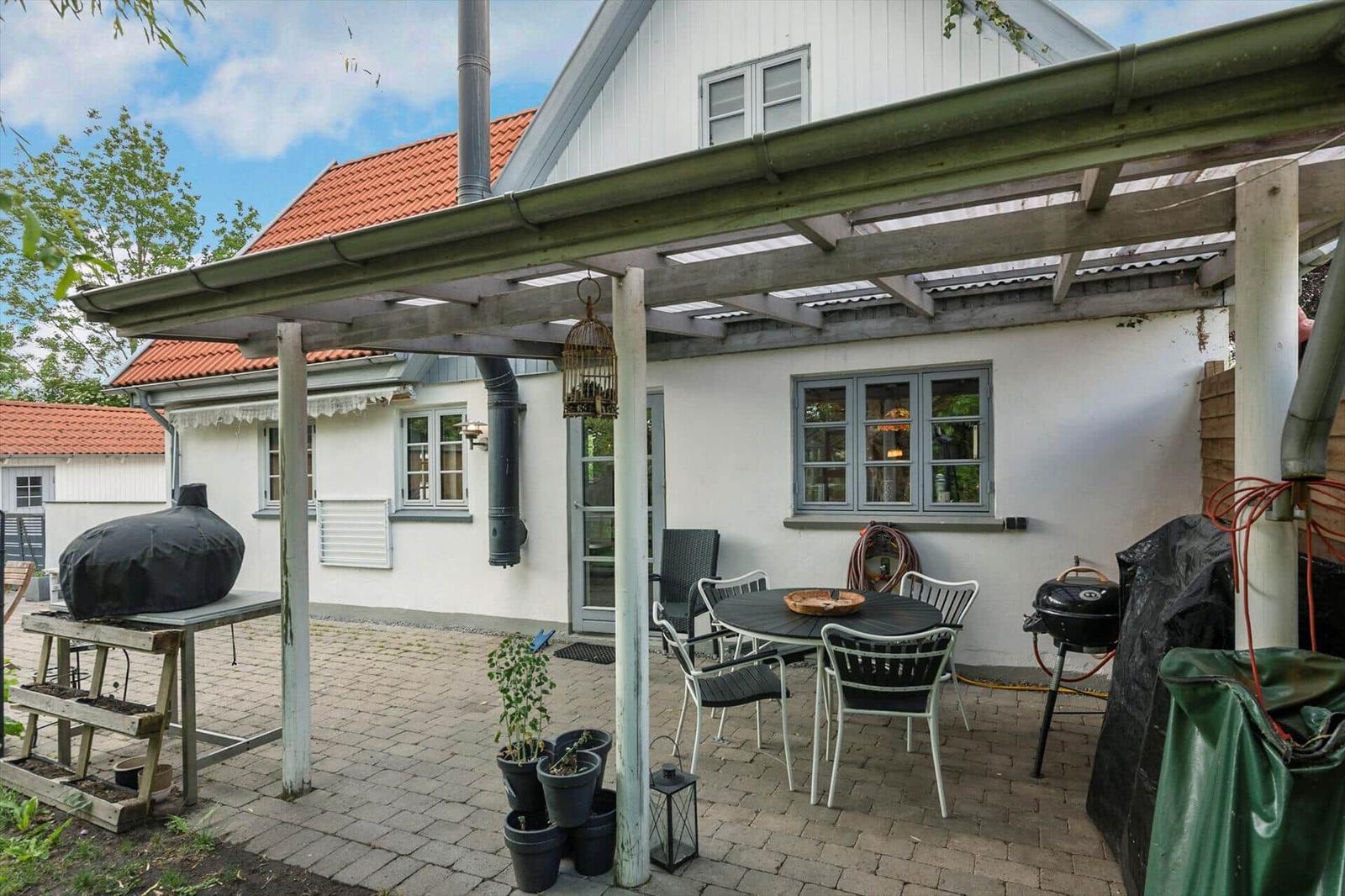Terrasse mit Tisch, Stühlen, Grill und Pergola unter einem blauen Himmel.