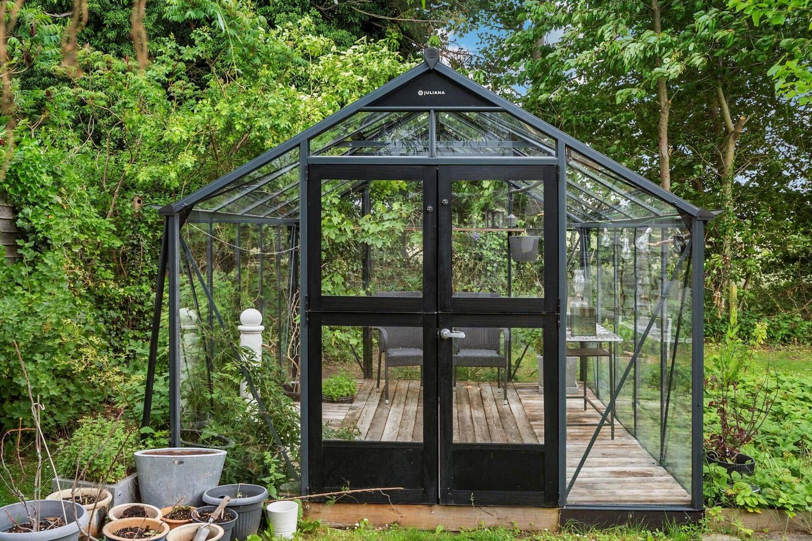 Gartenhaus mit Glaswänden und Holzboden. Im Inneren sind Stühle und Tisch sichtbar.