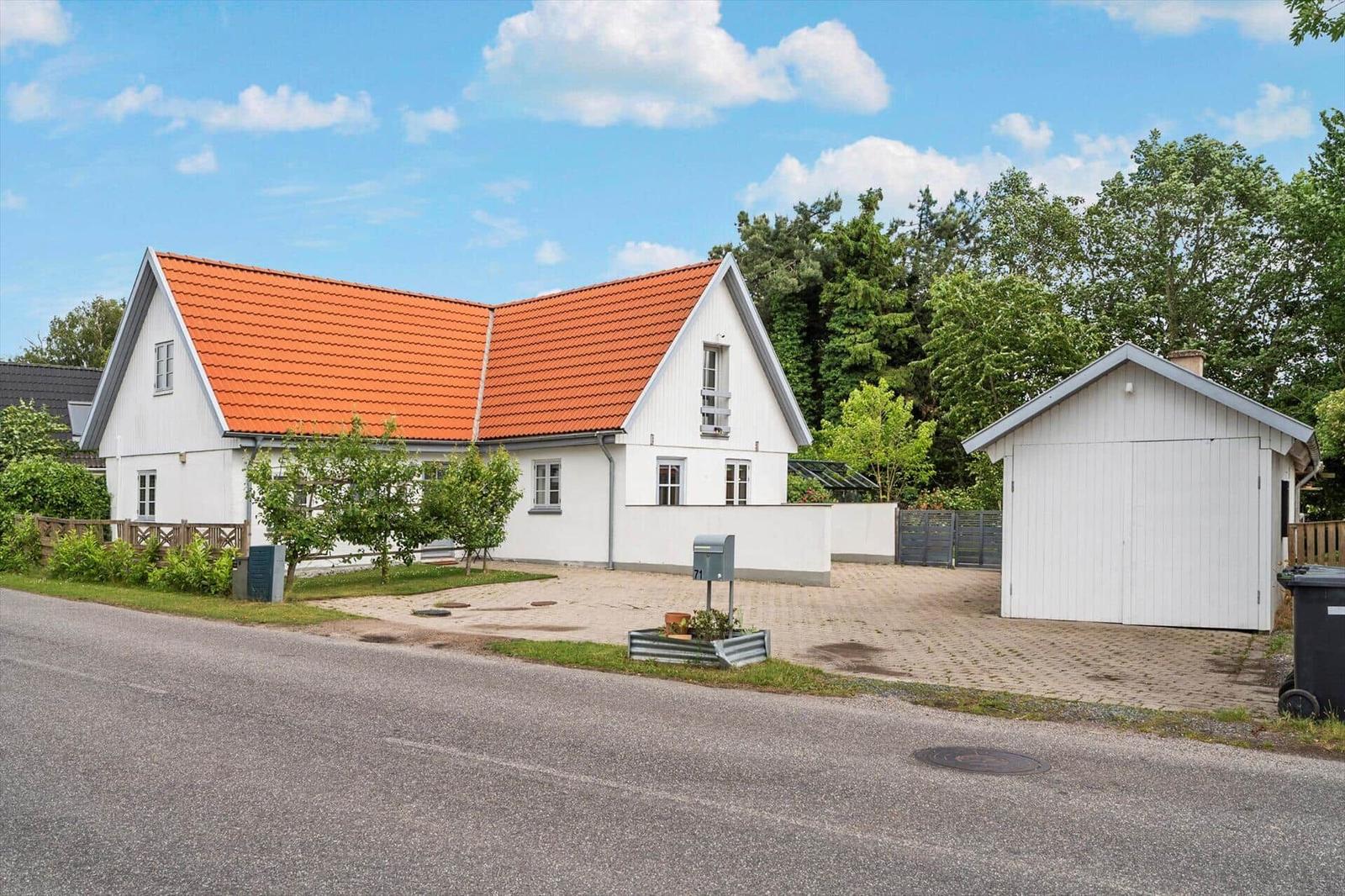 Weißes Haus mit rotem Dach und Garagenbereich. Grün umgeben und asphaltierter Vorplatz.