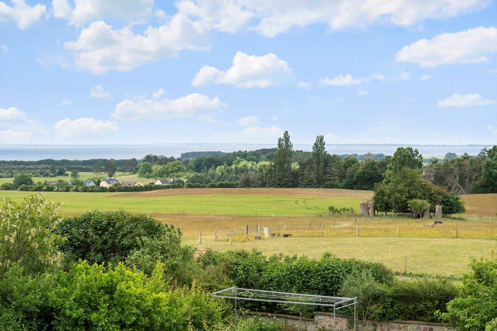 Weitläufiger Blick auf Felder, Bäume und das Meer unter blauem Himmel mit weißen Wolken.