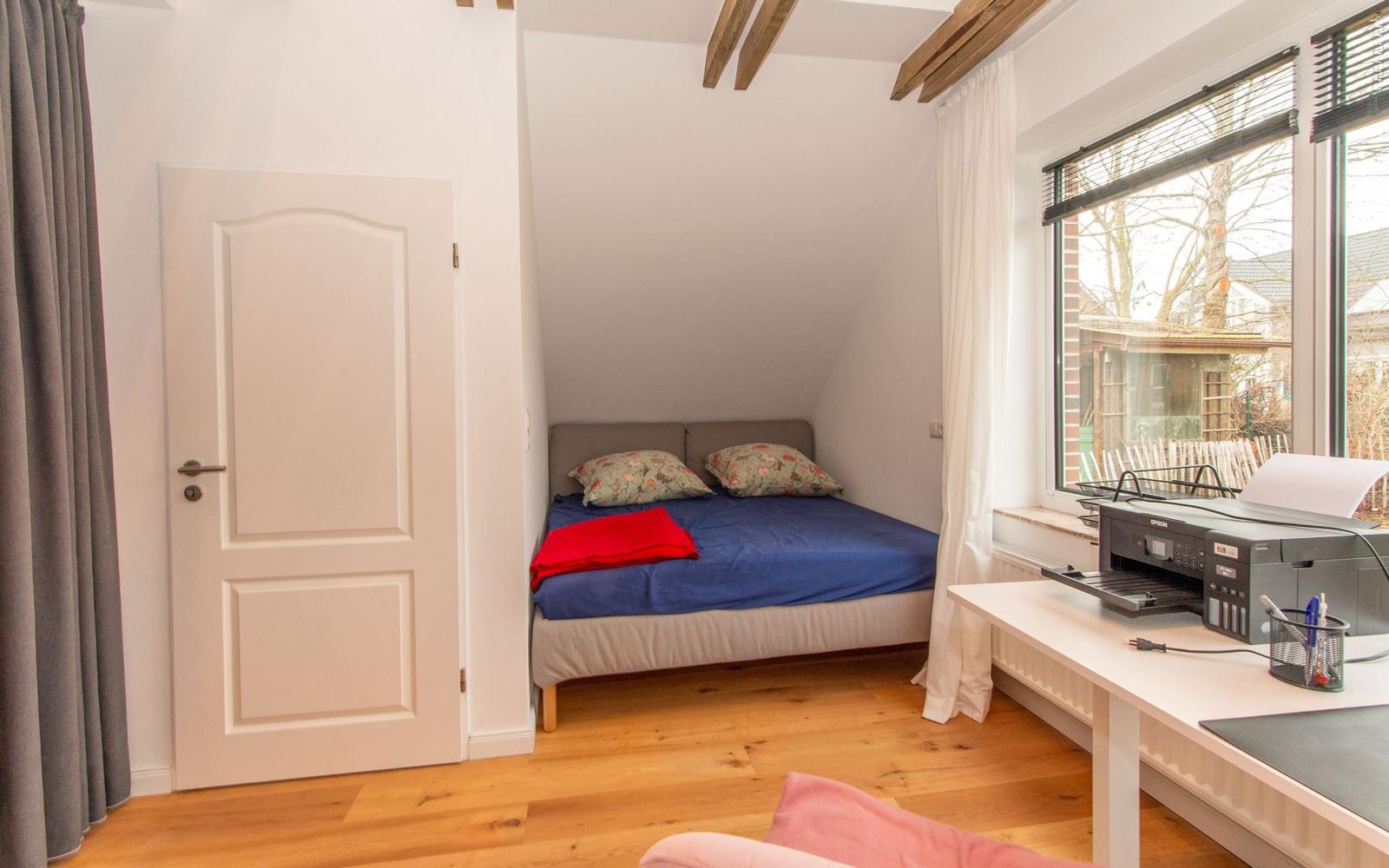 Schlafzimmer mit Bett, Schreibtisch und Fenster. Holzboden und Deckenbalken.