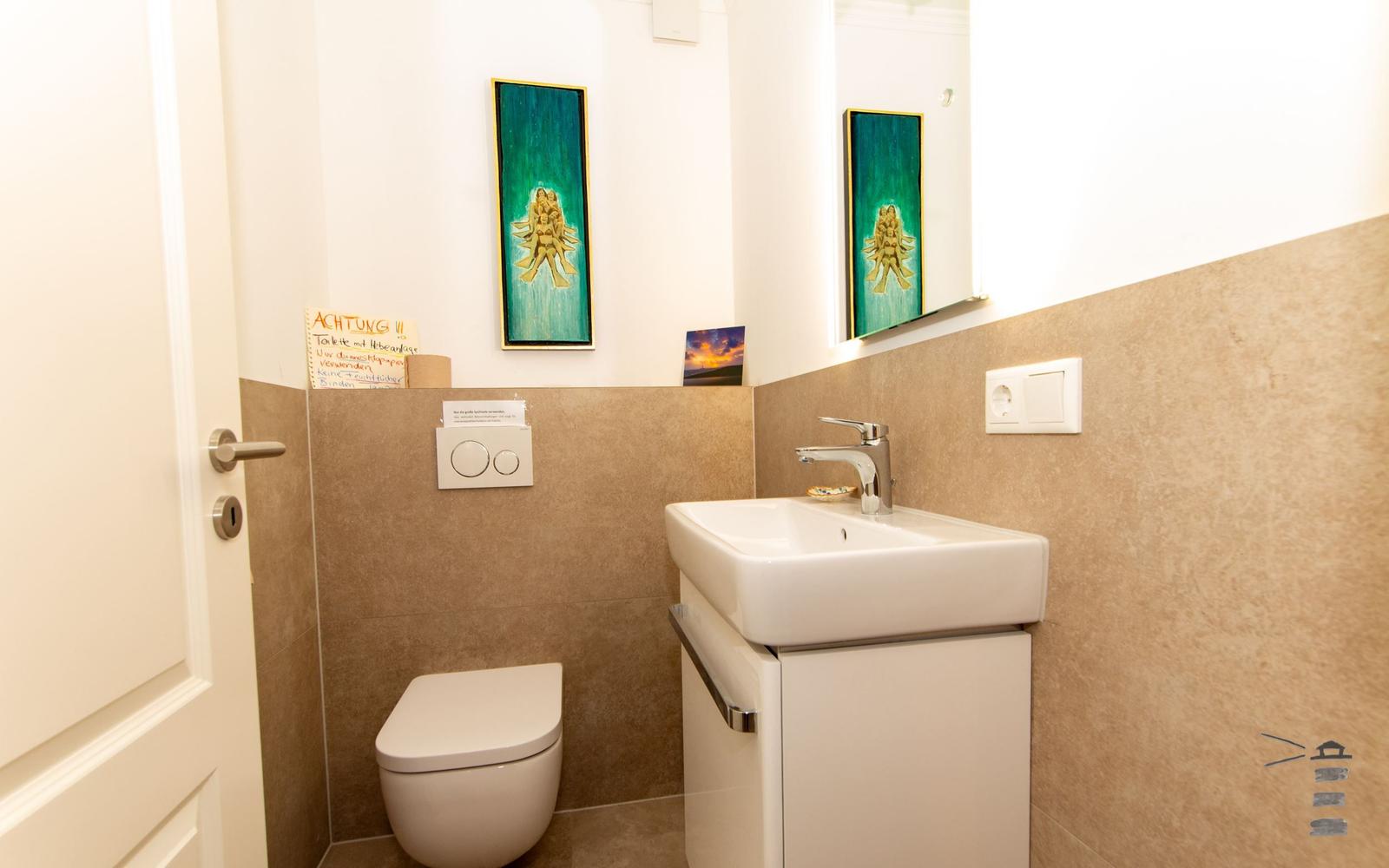 Badezimmer mit Toilette, Waschbecken und Spiegel. Wandbild und Hinweisschild sichtbar.