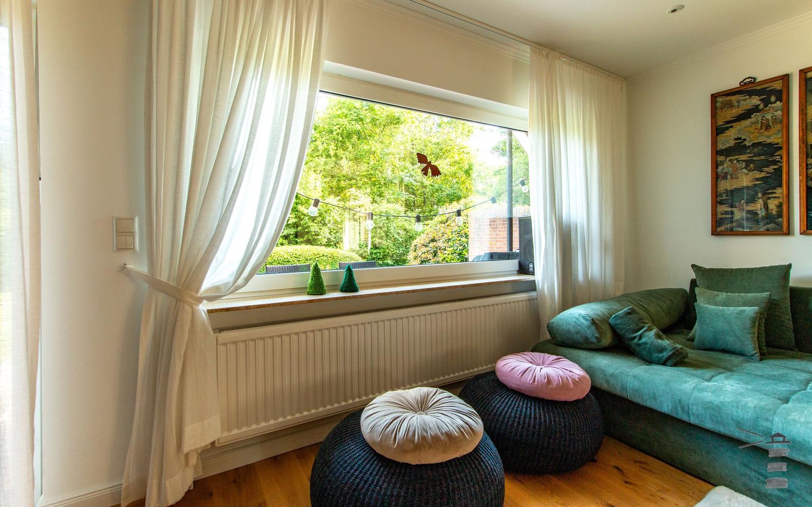 Ein Raum mit grünem Sofa, bunten Sitzkissen und großem Fenster mit Blick auf einen Garten.