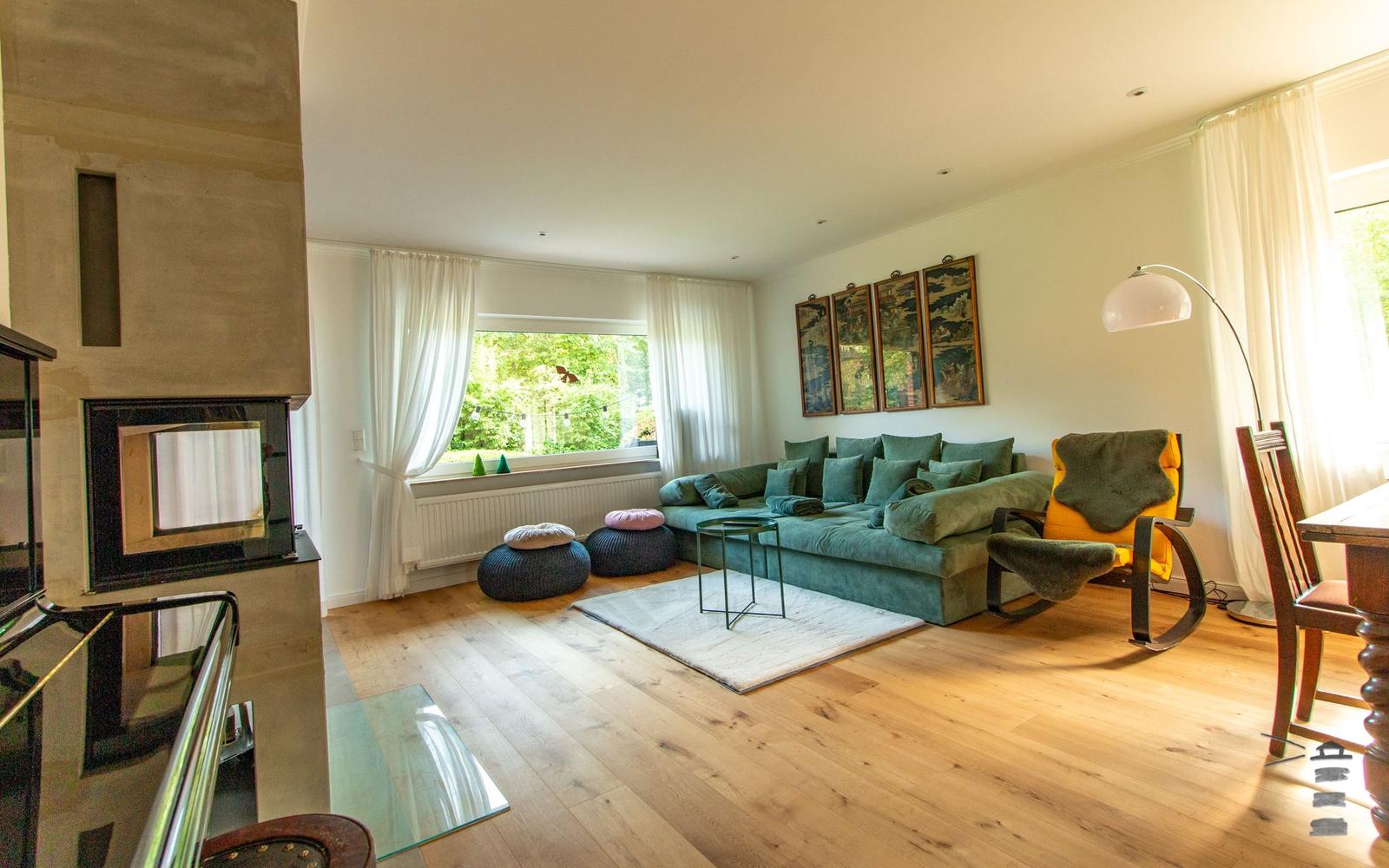 Wohnzimmer mit grünem Sofa, Holzboden und Kamin. Fenster mit Blick auf Garten.