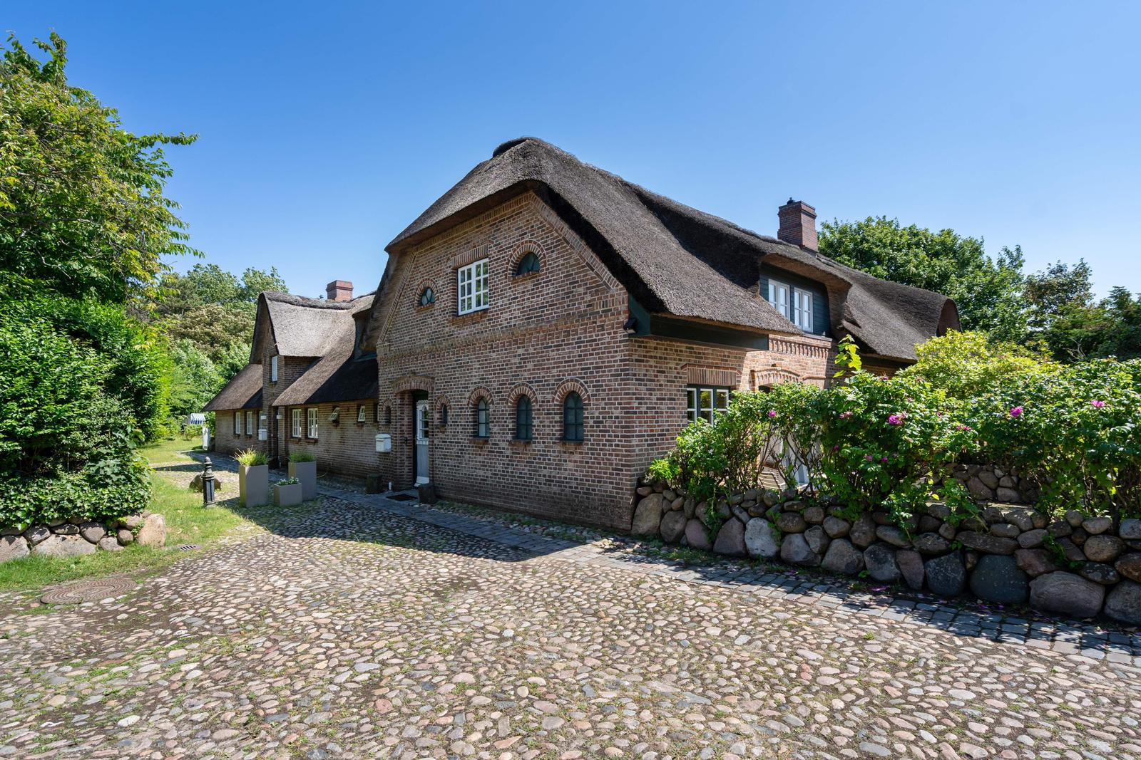 Ein traditionelles Haus mit Strohdach und Backsteinfassade, umgeben von Grün und Steinmauern.