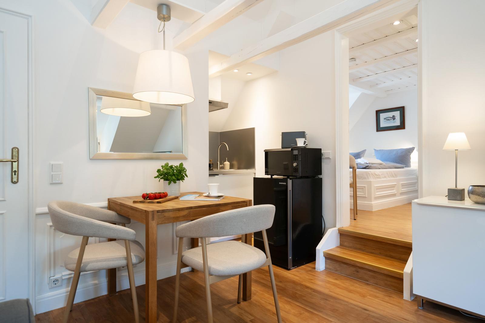 Essbereich mit Tisch und Stühlen, Küchenzeile und Schlafzimmer mit Bett im Hintergrund.