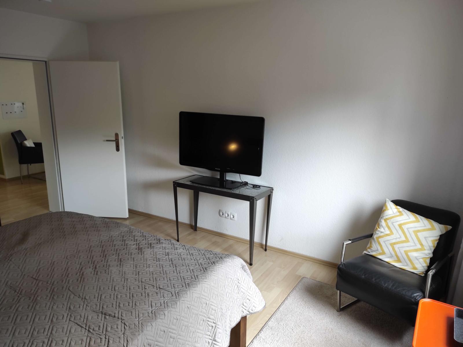 Schlafzimmer mit Bett, TV auf Tisch und Sessel mit Kissen.