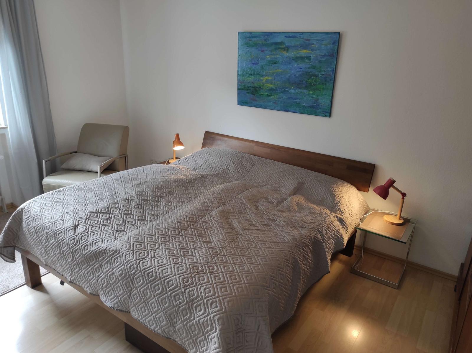 Schlafzimmer mit Bett, Nachttisch und Sessel. Wandbild und Lampen sichtbar.