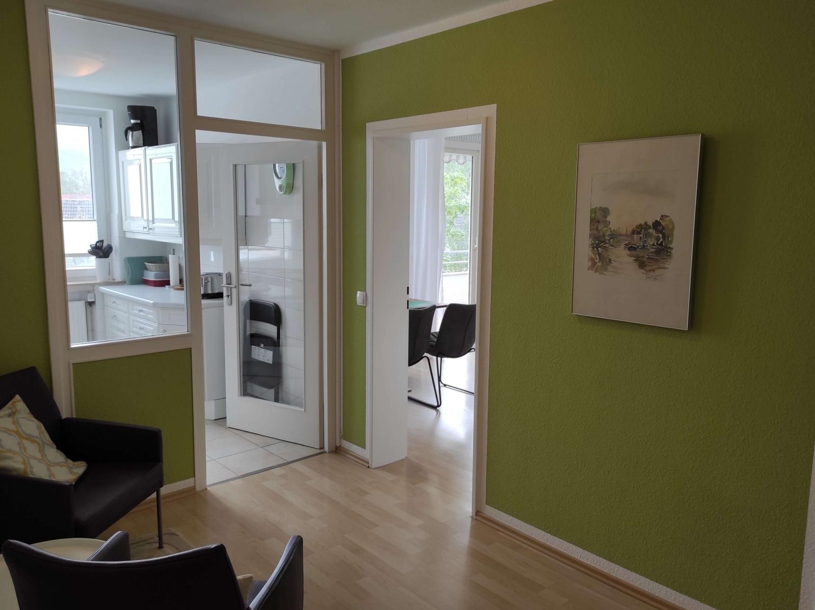 Wohnbereich mit grüner Wand, Sitzgelegenheiten und Blick in Küche und Esszimmer.
