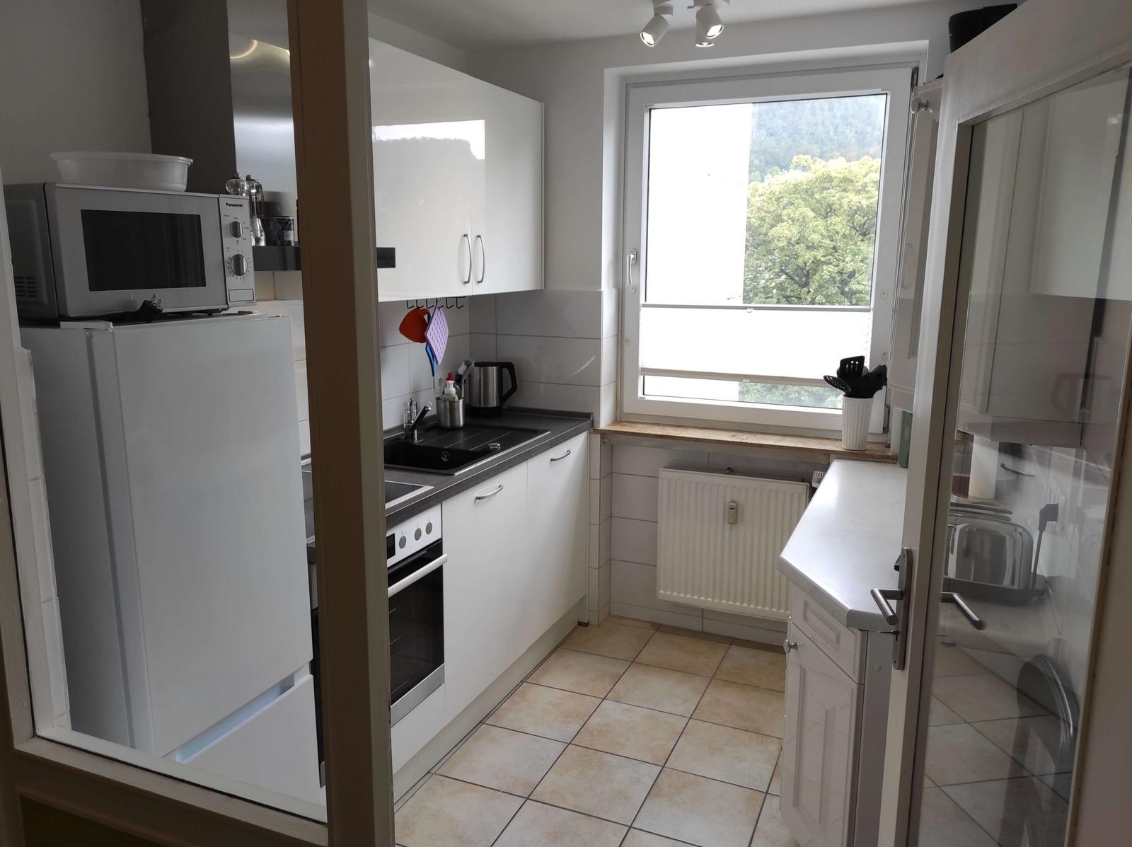 Küche mit weißen Schränken, Fenster mit Blick auf Wald, Kühlschrank, Mikrowelle und Herd.