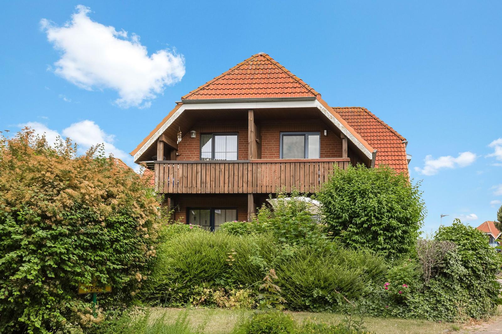A two-story house with a balcony and red roof, surrounded by green bushes.