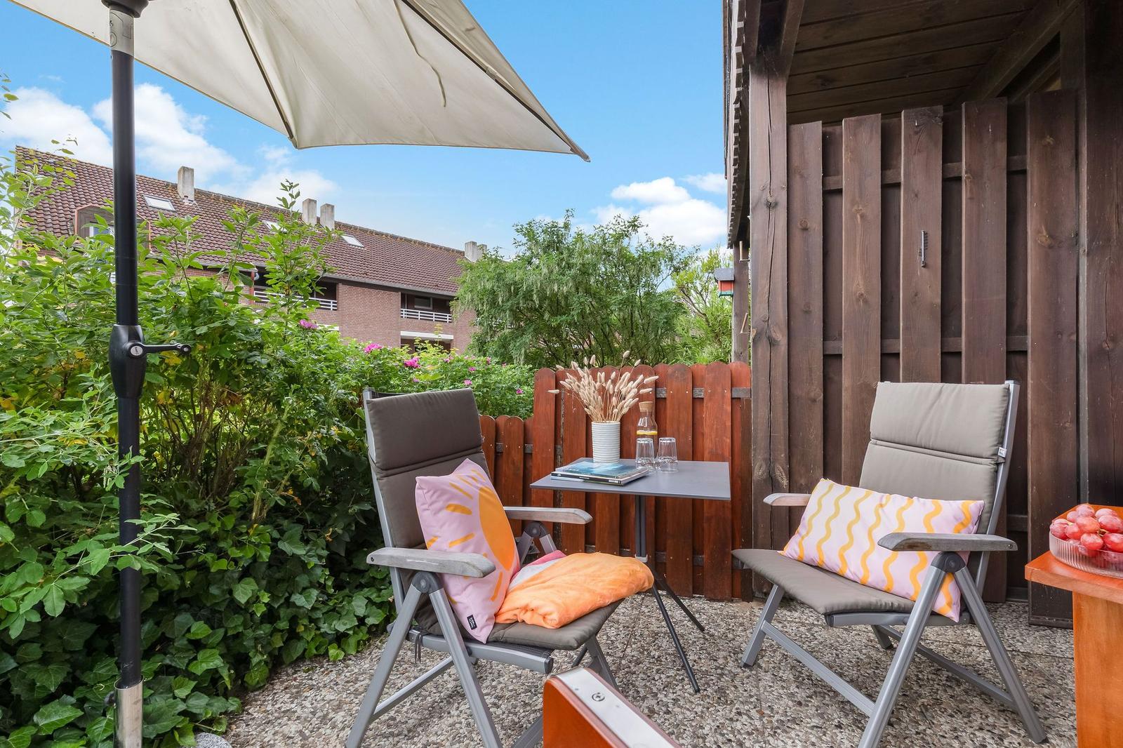 Terrace with two chairs, table, and umbrella. Background: green plants and wooden house.